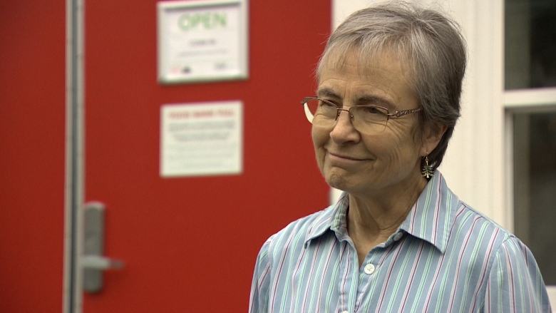 A woman with short, grey hair wears glasses and a striped button-up shirt. She stands in front of a bright red door. 