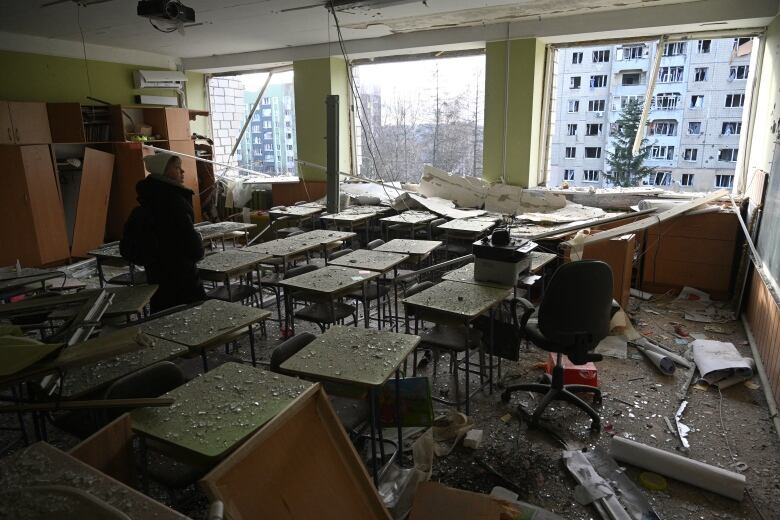 A woman stands in a classroom with three large windows, each of which has been blown out. Furniture is toppled and small glass shards are scattered.