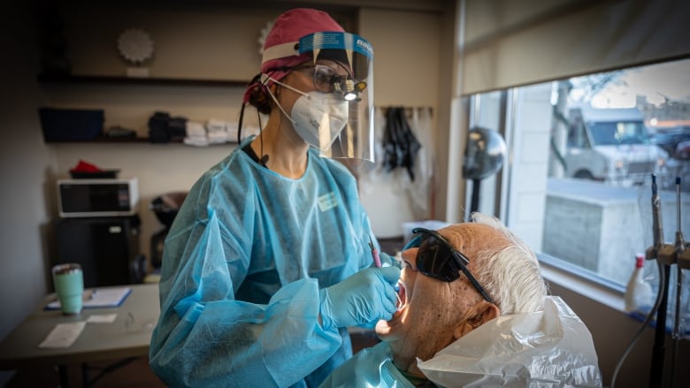 A dental hygienist dressed in scrubs, gloves and a mask peers inside the mouth of a patient doing an exam.