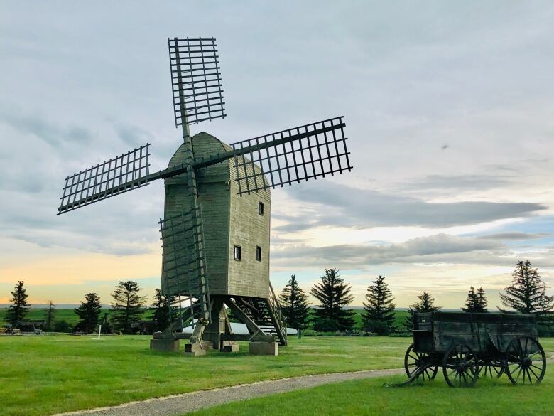 A windmill stands in a field with a covered wagon in front. 