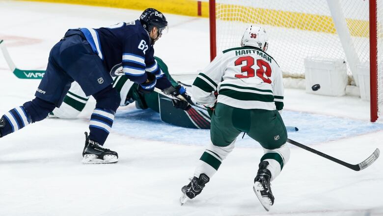 A hockey player fires a puck into the net, as a defender chases him.