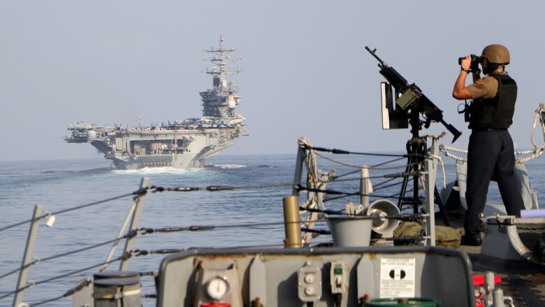 A U.S. sailor stands watch with binoculars on a ship sailing behind an aircraft carrier.