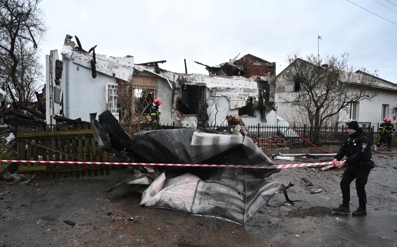 A police officer cordons off a piece of wreckage as firefighters inspect a damaged building.