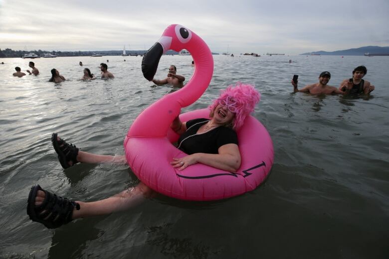 A woman floating in an inflatable pink flamingo, on beachwater, smiles for the camera. Behind her, other people also dip in the water.