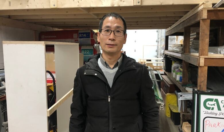 A man with glasses wearing a black coat stands in front of a wooden frame and shelves.