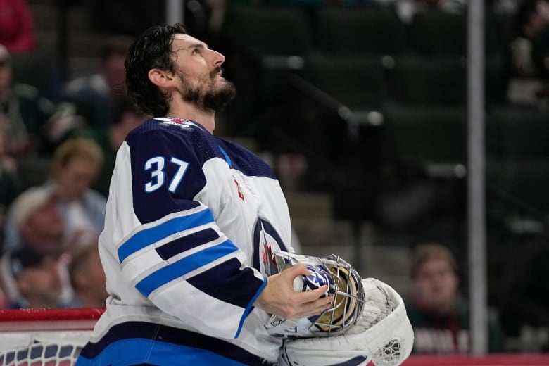 A hockey goalile holds his mask and looks up at the sky. He has black hair and a beard.