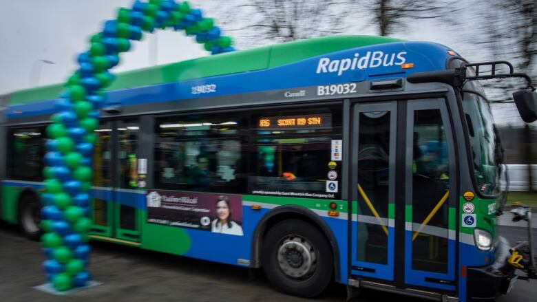 A green-and-blue bus with 'RapidBus' printed on the top is seen driving through an arc of green and blue balloons. 