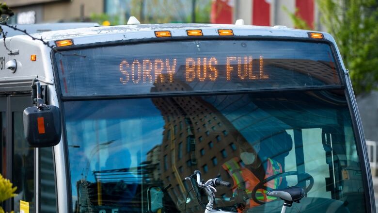 A bus driver wearing a high-vis vest and a mask waits at a stop, with a sign above him reading, 'Sorry Bus Full.'