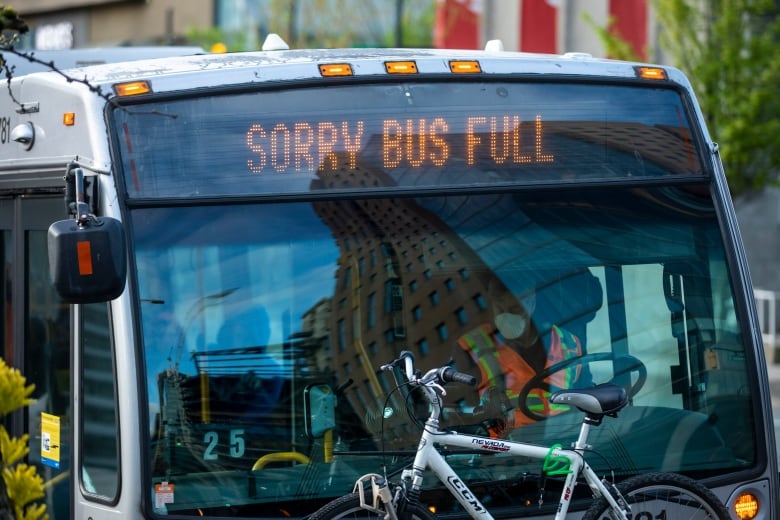 A bus driver wearing a high-vis vest and a mask waits at a stop, with a sign above him reading, 'Sorry Bus Full.'