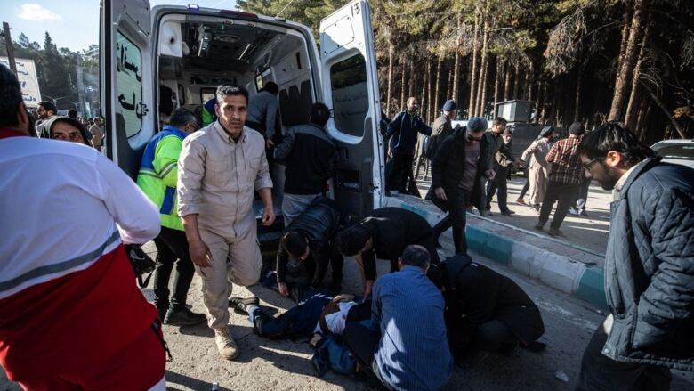 A group of people crowd around a person prone on the ground behind an ambulance with its back doors open.