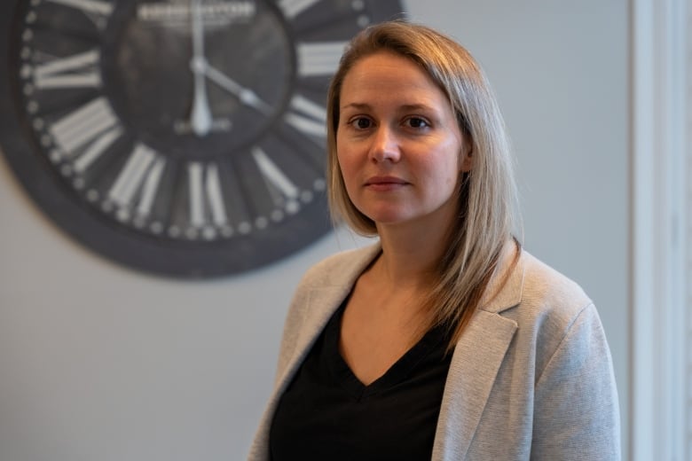 A woman with blond hair and wearing a beige blazer over a black top stands in an indoor room, with a large clock seen behind her.