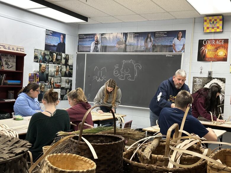 A classroom full of students shows them sitting at tables weaving strands of wood into baskets.