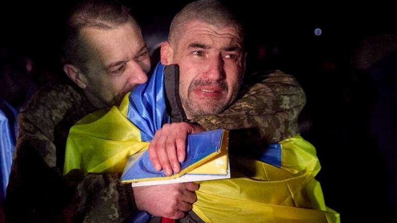 A man wearing a military uniform embraces another man wrapped in a Ukrainian flag.