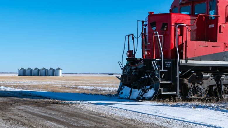 A photo shows light snow on a field with grain bins in the background and the front of a train in the foreground.