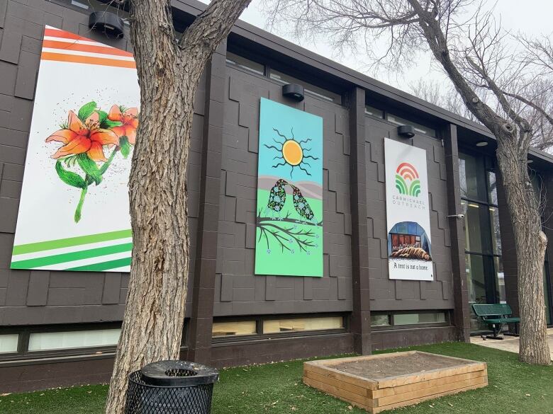 Bright, colourful Indigenous paintings line a brown wall. Trees line the wall as well. 