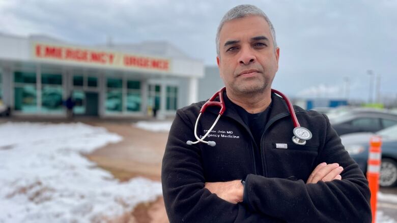 A man in a dark coat and a stethoscope around his neck stands in front of an emergency department.