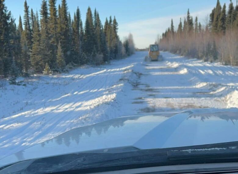 A piece of machinery can be seen in the distance working on a snow-covered road surrounded by trees in a photo taken from inside another vehilcle.