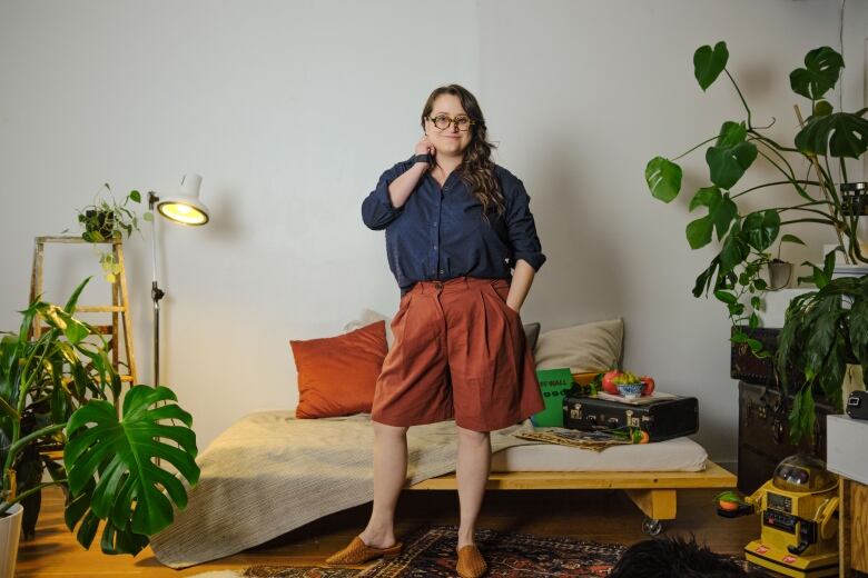 A woman is wearing a blue button up shirt and orange shorts. Behind her is a couch with beige and white cushions and plants on either side. 