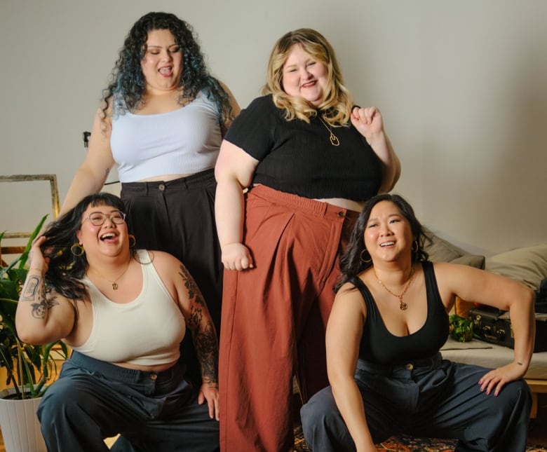 Four women are smiling and laughing in front of a black fan. Two of the women are standing and the other two are crouching in front. They are all wearing size inclusive clothing. 