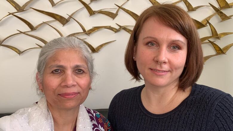 An elderly woman sits beside a younger woman with chin-length hair in front of a bird motif.