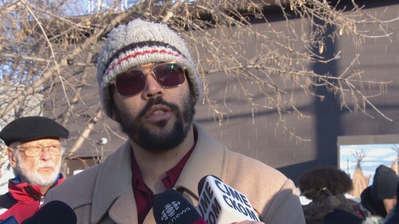 A man in a toque and sunglasses speaks into microphones. 