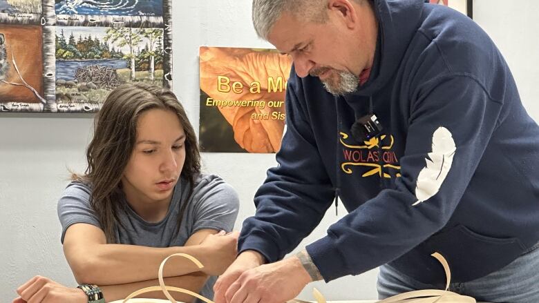 A man with grey hair and beard leans over a table, showing a teenager with long dark hair how to weave a basket.