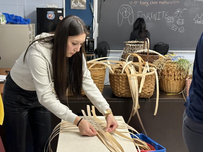 A girl with long dark hair bends over a table, weaving together strands of wood into a basket.