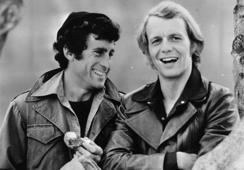 Two men, one at left with dark hair and one at right with blond hair, stand outdoors laughing, wearing in jackets with large, spread collars.