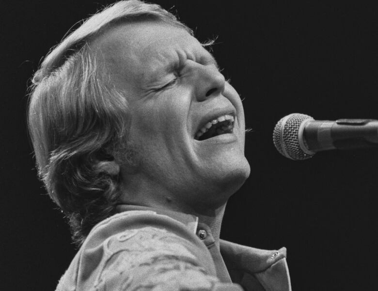 Close-up image of a man with his eyes closed, singing into a microphone and playing a guitar.