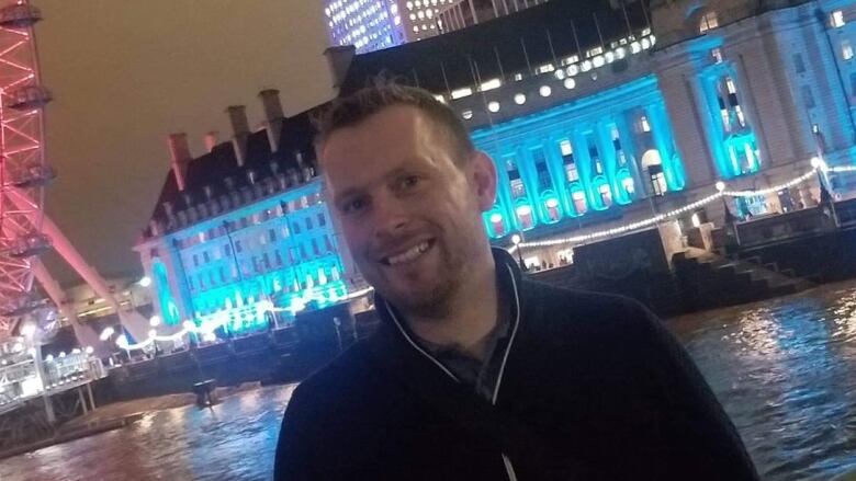 A man smiles on a bridge over a river, with an old building lit up behind him.