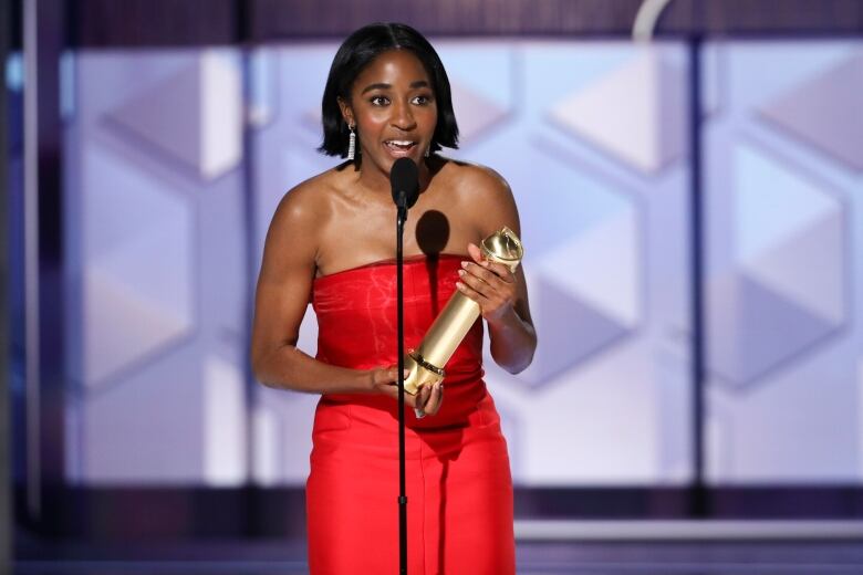 A woman wearing a bright red dress speaks into a microphone while holding a golden statuette with both hands.