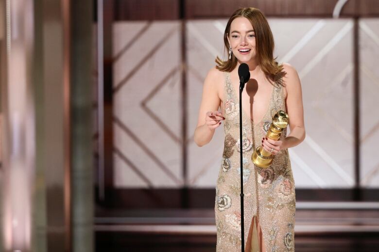 A woman wearing a gown printed with flowers speaks into a microphone while holding a golden statuette with one hand.