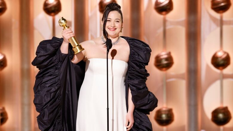 A woman wearing a white gown and a black shawl holds a golden statuette in one hand while speaking into a microphone.