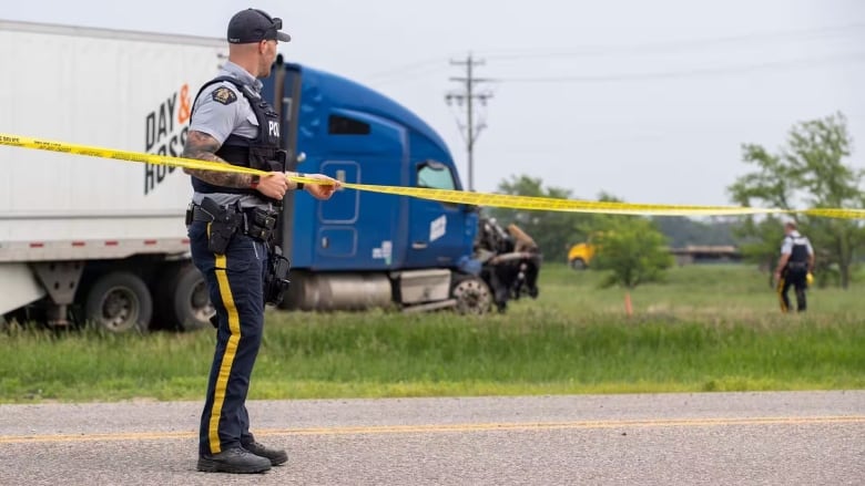 RCMP officers string out yellow tape across a highway after a crash. A semi-truck with a damaged front can be seen in the background.