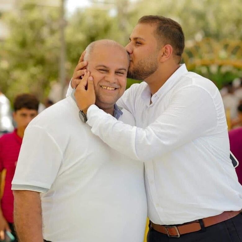 A young man wraps his arms around an older man's head and plants a kiss on his temple. The older man smiles brightly at the camera.