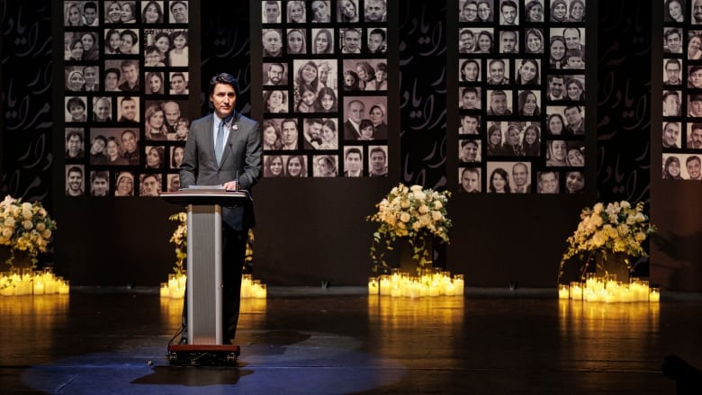 A man in a suit speaks at a podium in front of a wall commemorating victims of Flight PS752.