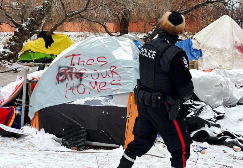 An RCMP officer in winter uniform looks at a cluster of tents. One is painted with the words, 