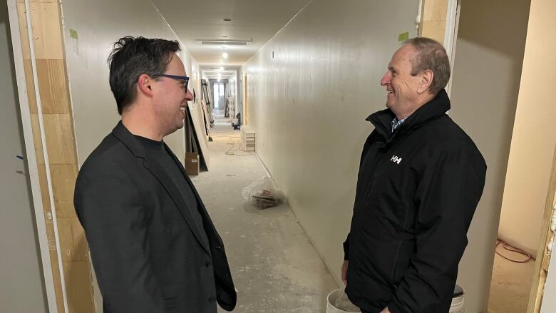 Dan Kutcher and Bobby Morrissey stand facing each other smiling. The building they are in and hallway are under construction. 