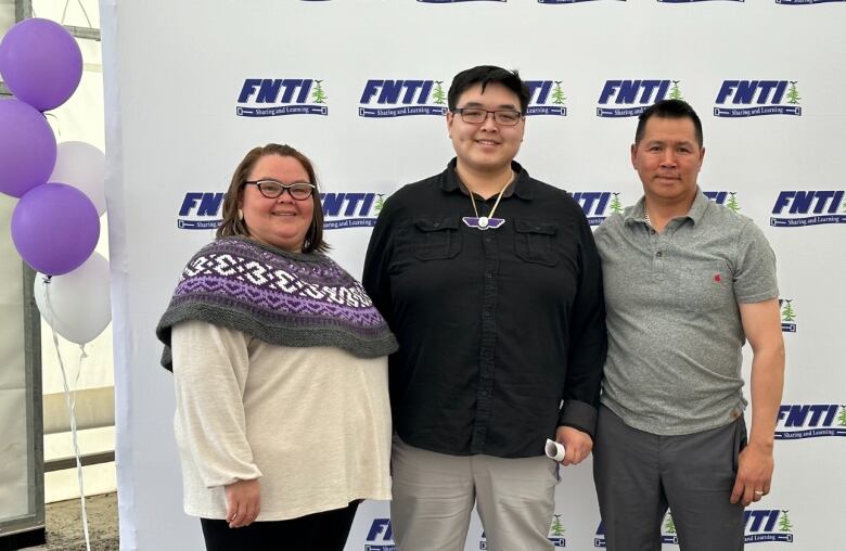 A young man stands flanked by a mother and father in front of a panel imprinted with a school's logo