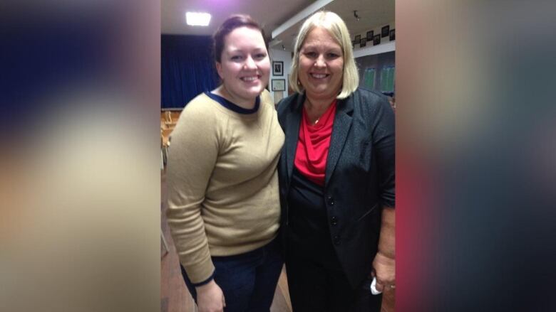 Two women stand and smile at a camera. 