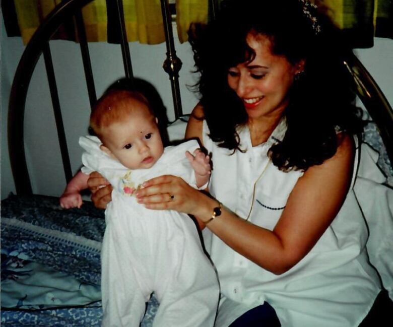 A smiling woman with long dark curly hair sitting on a bed and holding a baby.