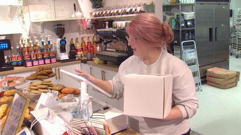 A woman with long pink hair holds a cake box as she uses a cash register in a pink bakery.