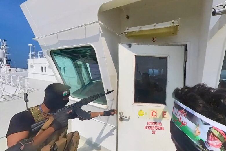 A man wearing a black head covering and a khaki flak jacket points an automatic weapon into the cabin of a ship. 