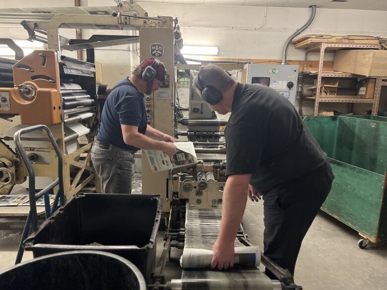 Two men wearing protective earphones handle newspapers as they come off a printing press.