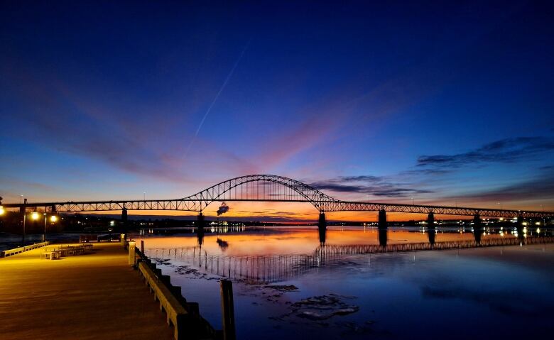 A bridge over blue water, silhouetted black against an orange sunset.