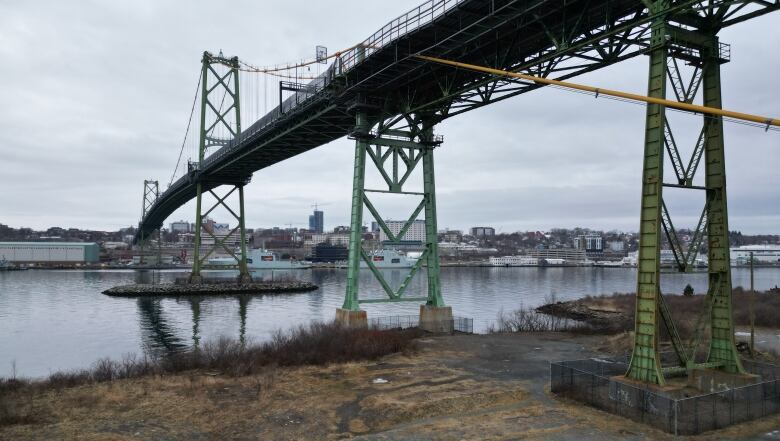 A suspension bridge is seen over the harbour on an overcast day. The closest metal footing under the bridge is patchy with rust, while the one beside it is a lighter green