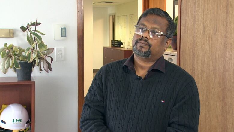 A Bangladeshi man with glasses and a black sweater stands in an office with a brown door open behind him