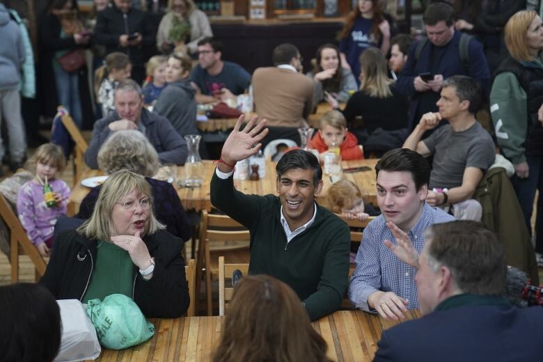 A man wearing a sweater smiles and raises his right arms while sitting at a table alongside several other adults.