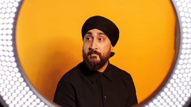 A man wearing a turban poses for a portrait in front of an orange backdrop with a ring light framing his face.