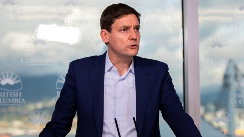 A white man wearing a light blue shirt and a darker blue coat speaks at a podium.